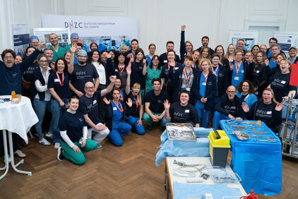 Eine große Gruppe von Menschen steht zusammen und lächelt, einige heben die Hände. Alle tragen einheitliche T-Shirts mit einem Logo. Im Hintergrund sind Informationsstände und medizinische Ausrüstung zu sehen. Die Atmosphäre wirkt freundlich und einladend.