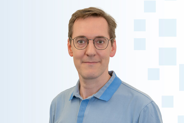 Ein Mann mit Brille und kurzem, braunem Haar trägt ein hellblaues Poloshirt. Er lächelt leicht und steht vor einem hellen Hintergrund mit geometrischen Mustern.