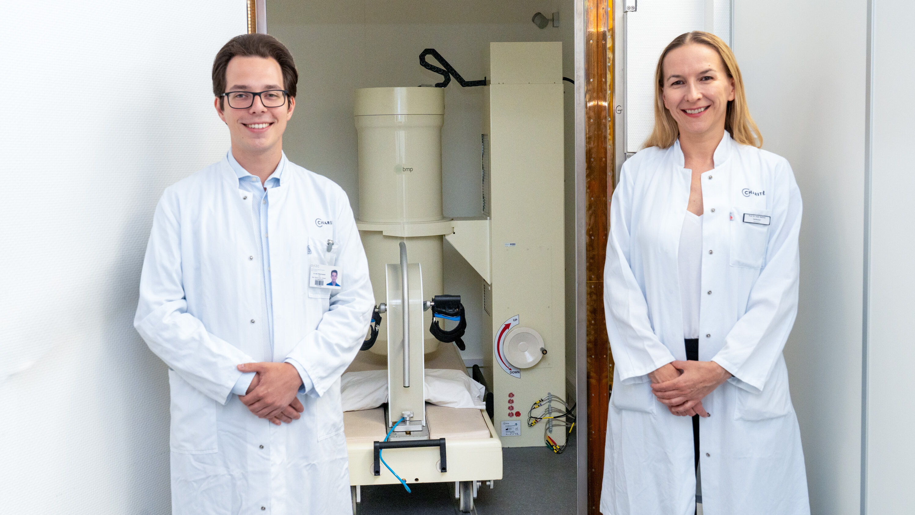 Zwei Ärzte, ein Mann und eine Frau, stehen lächelnd vor einem medizinischen Gerät in einem Klinikraum. Beide tragen weiße Laborkittel. Das Gerät befindet sich hinter ihnen und scheint für diagnostische Zwecke verwendet zu werden.