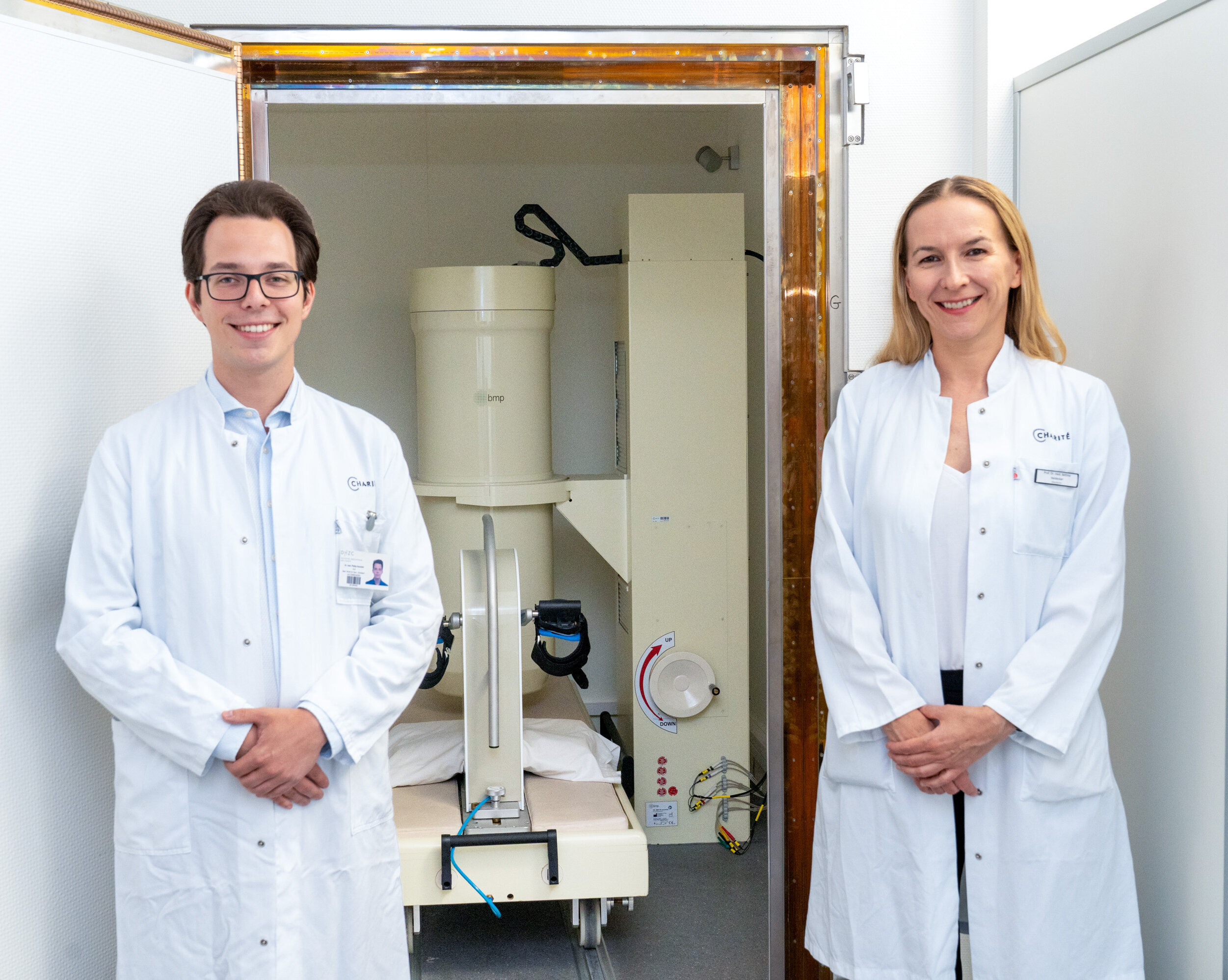 Zwei Ärzte, ein Mann und eine Frau, stehen lächelnd vor einem medizinischen Gerät in einem Klinikraum. Beide tragen weiße Laborkittel. Das Gerät befindet sich hinter ihnen und scheint für diagnostische Zwecke verwendet zu werden.