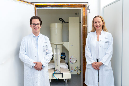 Zwei Ärzte, ein Mann und eine Frau, stehen lächelnd vor einem medizinischen Gerät in einem Klinikraum. Beide tragen weiße Laborkittel. Das Gerät befindet sich hinter ihnen und scheint für diagnostische Zwecke verwendet zu werden.
