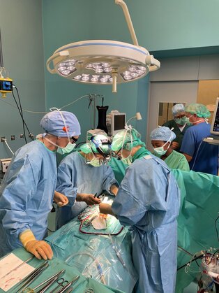 Eine Gruppe von Chirurgen und medizinischem Personal steht während einer Operation um einen Patienten, der auf einem Tisch liegt. Alle tragen OP-Kleidung, Masken und Hauben. Die Umgebung ist steril, mit chirurgischen Instrumenten und Beleuchtung über dem Operationstisch.