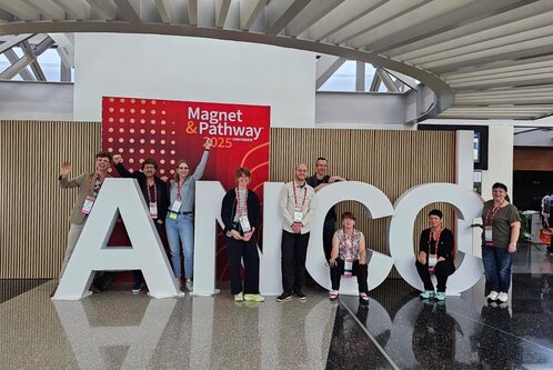 Eine Gruppe von acht Personen posiert fröhlich vor großen, weißen Buchstaben, die das Wort "AHA" bilden. Im Hintergrund steht ein Banner zur Konferenz "Magnet & Pathway 2025". Die Atmosphäre ist festlich und einladend.