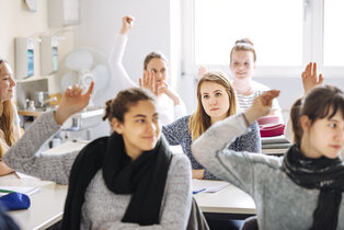 Eine Gruppe von Schülerinnen sitzt in einem Klassenzimmer. Einige haben ihre Hände gehoben, um sich zu melden, während eine Schülerin in der Mitte aufmerksam in die Kamera schaut. Das Bild vermittelt eine Lernumgebung, in der aktive Teilnahme gefördert wird.
