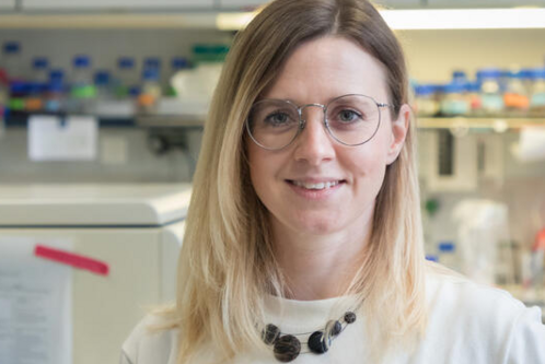 Eine lächelnde Frau mit langen, blonden Haaren und Brille steht in einem Labor. Sie trägt ein helles Shirt und eine auffällige Halskette. Im Hintergrund sind Laborutensilien und Flaschen sichtbar, die auf eine wissenschaftliche Umgebung hinweisen.