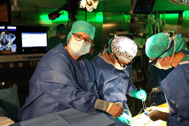 In einem Operationssaal arbeiten mehrere medizinische Fachkräfte zusammen. Eine Ärztin mit blauer OP-Bekleidung und einer Schutzmaske blickt in die Kamera. Zwei weitere Fachkräfte sind beschäftigt, während sie chirurgische Eingriffe durchführen, und das Licht ist fokussiert auf den Operationstisch.