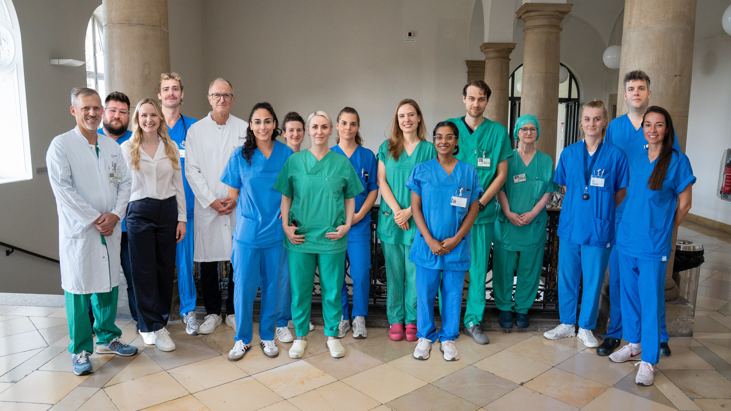 Eine Gruppe von 15 Personen steht in einem Innenraum. Sie tragen verschiedene medizinische Uniformen in Blau und Grün sowie weiße Kittel. Die Personen lächeln und wirken freundlich, vor einer neutralen Hintergrundarchitektur mit Säulen.