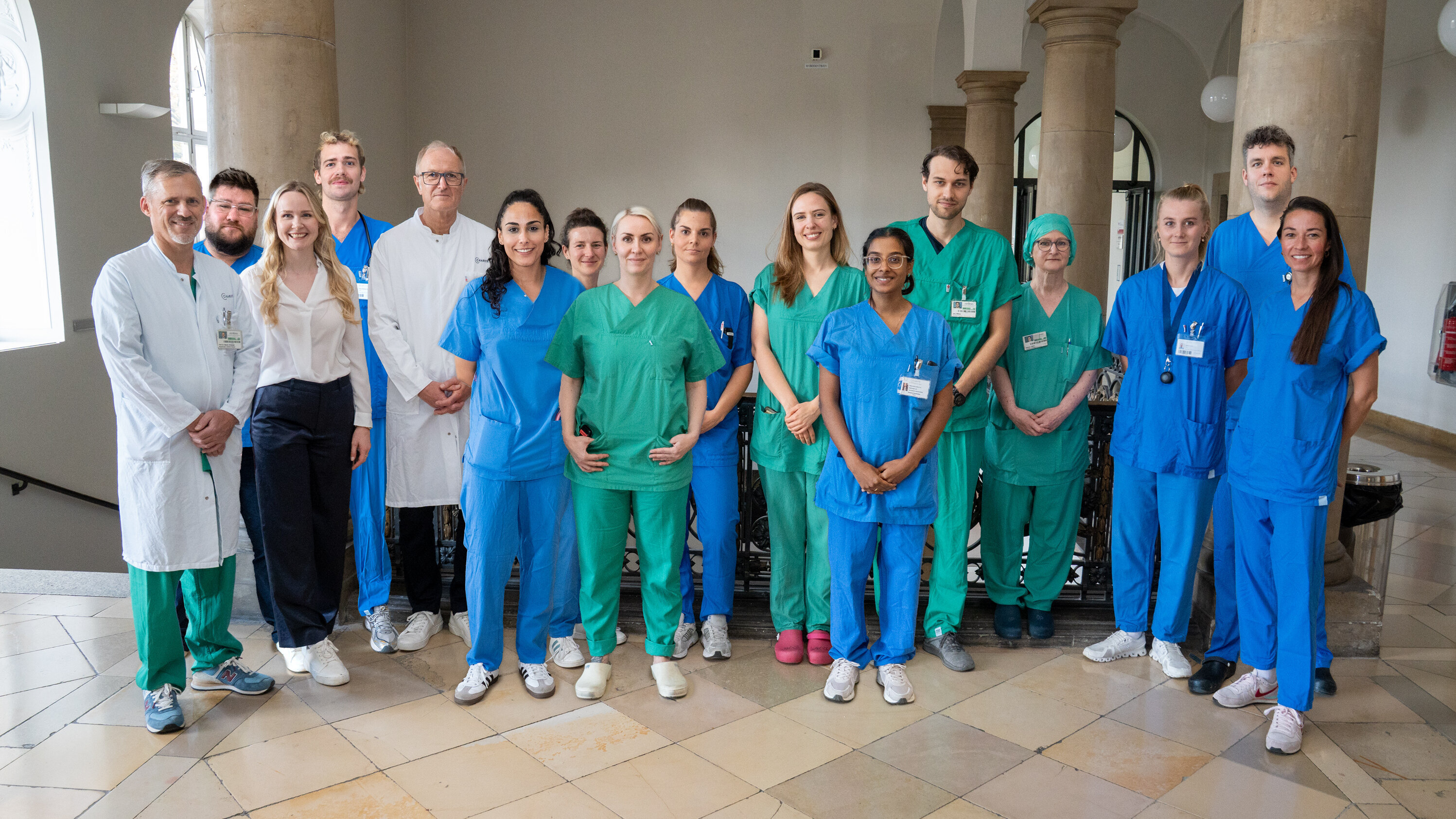 Eine Gruppe von 15 Personen steht in einem Innenraum. Sie tragen verschiedene medizinische Uniformen in Blau und Grün sowie weiße Kittel. Die Personen lächeln und wirken freundlich, vor einer neutralen Hintergrundarchitektur mit Säulen.