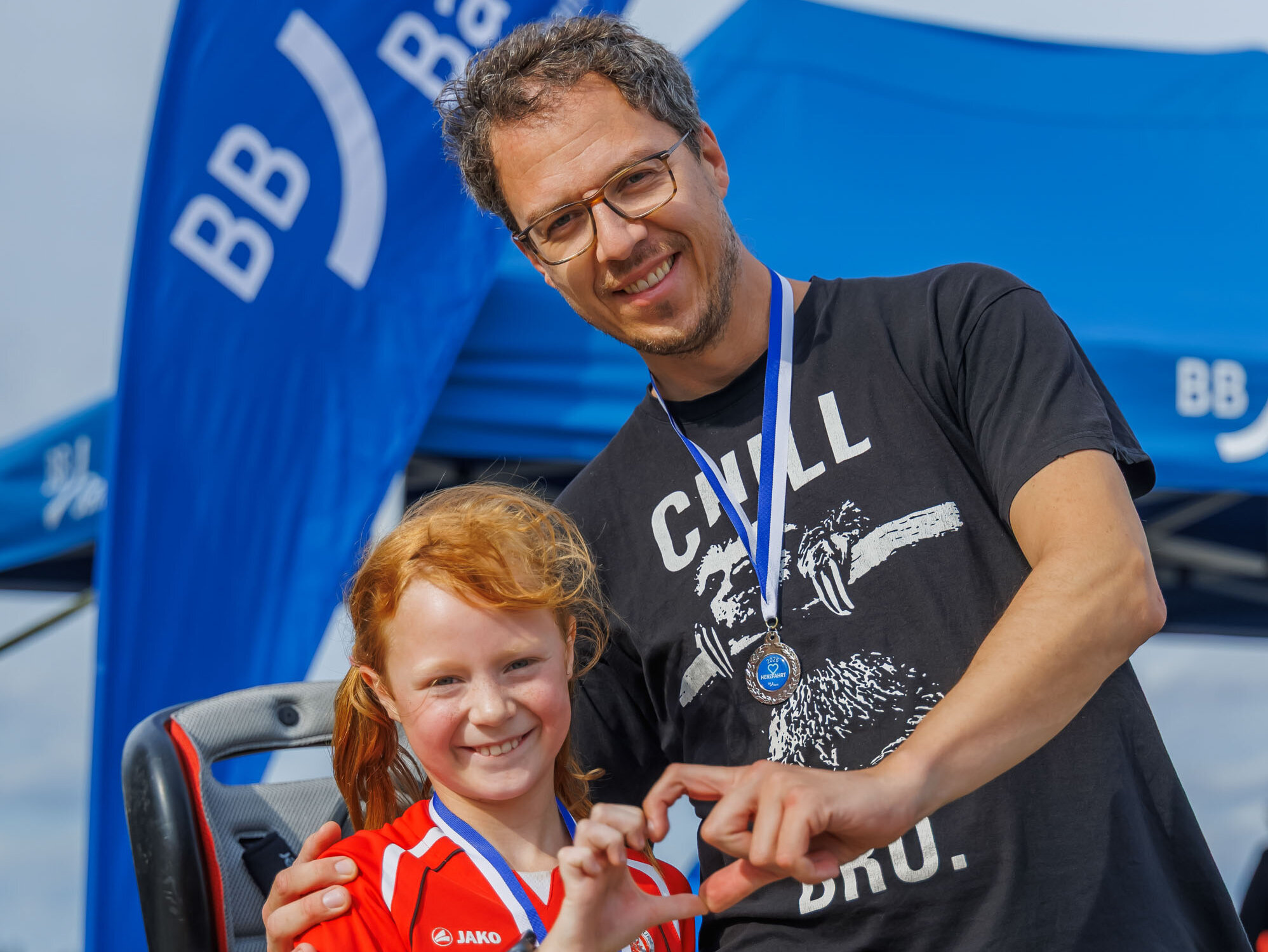 Ein Mann und ein Mädchen stehen lächelnd nebeneinander. Beide tragen Medaillen. Das Mädchen in einem roten Sport-Shirt zeigt mit ihrer Hand ein Herzzeichen. Im Hintergrund sind Zelte und Fahnen sichtbar. Die Atmosphäre ist festlich und unterstützend.