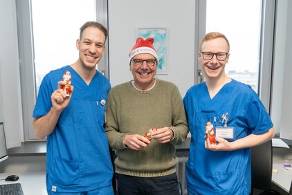 Drei Männer in blauen Arztkitteln stehen lächelnd in einem Büro. Der Mann in der Mitte trägt einen Weihnachtsmann-Hut und hält eine Schokoladenfigur eines Weihnachtsmanns in der Hand. Die Männer wirken freundlich und feiern offensichtlich die Weihnachtszeit.