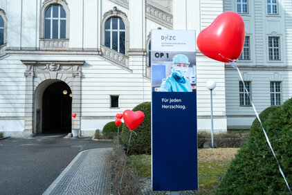 Das Bild zeigt ein Schild mit der Aufschrift "Für jeden Herzschlag", umgeben von roten Herzluftballons. Im Hintergrund ist ein Gebäude mit großen Fenstern zu sehen. Der Fokus liegt auf der Botschaft, die Herzgesundheit zu fördern.
