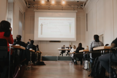 Eine Präsentation in einem Seminarraum wird gezeigt. Im Vordergrund sind Teilnehmer zu sehen, die auf einen Bildschirm schauen. Der Bildschirm zeigt Diagramme und Informationen zu einem medizinischen Thema an, während Referenten vor einem Tisch sitzen.