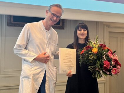 Ein lächelnder Mann im weißen Laborkittel steht neben einer Frau in schwarzem Kleid. Sie hält ein Zertifikat in der einen Hand und einen großen Blumenstrauß in der anderen. Beide scheinen sich über einen besonderen Anlass zu freuen.
