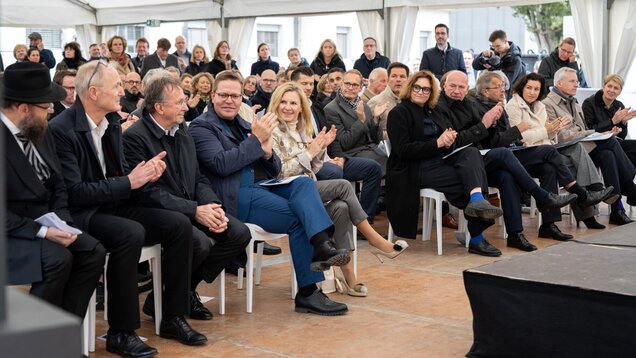 Eine Gruppe von Menschen sitzt in einem Zelt und applaudiert. Die Teilnehmer sind im Geschäftsstil gekleidet und zeigen unterschiedliche Emotionen von Freude bis Aufmerksamkeit. Der Hintergrund zeigt weitere ansässige Personen, die ebenfalls dem Geschehen folgen.