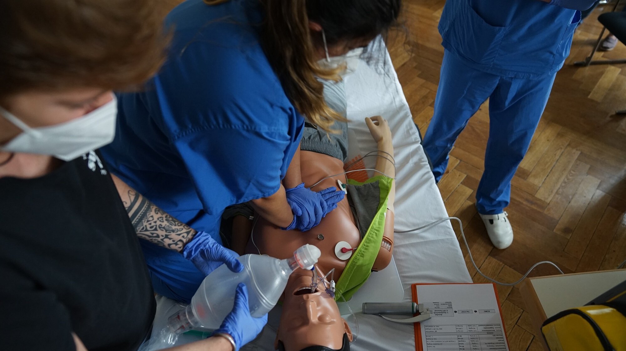 Zwei Personen in blauen Kitteln führen eine Erste-Hilfe-Maßnahme an einer menschlichen Puppe durch. Eine Person hält eine Beatmungsmaske auf das Gesicht der Puppe, während die andere bereitsteht, um Unterstützung zu leisten. Die Szene zeigt eine praktische Übung zur Wiederbelebung.