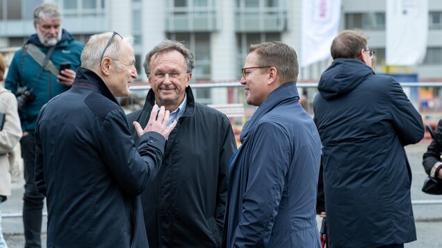 Drei Männer in dunklen Jacken stehen im Freien und unterhalten sich angeregt. Einer gestikuliert mit der Hand, während die anderen beiden aufmerksam zuhören. Im Hintergrund sind weitere Personen und eine städtische Umgebung zu sehen.