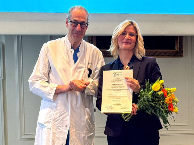 Ein Arzt in einem weißen Kittel steht neben einer Frau in einem dunklen Blazer, die ein Zertifikat und einen Blumenstrauß hält. Beide lachen und wirken erfreut. Der Hintergrund ist hell und professionell, was auf eine feierliche Veranstaltung hinweist.