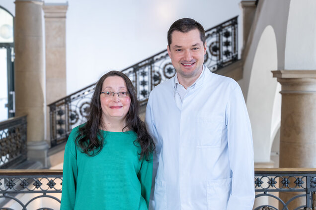 Zwei Personen stehen lächelnd nebeneinander in einem Innenraum. Die Frau trägt ein grünes Oberteil und hat lange, dunkle Haare. Der Mann trägt einen weißen Laborkittel. Im Hintergrund ist eine elegante Treppe und eine balustradierte Brüstung zu sehen.