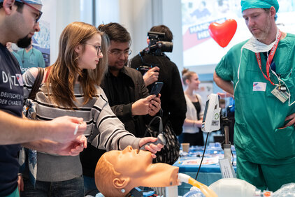Eine Gruppe von Menschen steht um einen Tisch mit medizinischen Geräten. Eine Frau mit langen Haaren und Brille hält einen Stethoskop an ein Modellkopf, während andere Zuschauer interessiert zuschauen. Ein Arzt in grüner Uniform erklärt die Geräte und deren Anwendung.