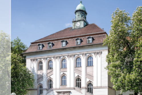 Ein historisches Gebäude mit einer weißen Fassade und großen Fenstern. Auf dem Dach befindet sich ein Uhrenturm mit einer Kupferkuppel. Rund um das Gebäude stehen einige Bäume, die zur Landschaft beitragen.