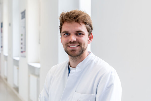 Ein junger Mann mit einem freundlichen Lächeln trägt einen weißen Arztkittel. Er steht in einem hellen Flur mit großen Fenstern im Hintergrund. Der Fokus liegt auf seinem Gesicht und seiner positiven Ausstrahlung.