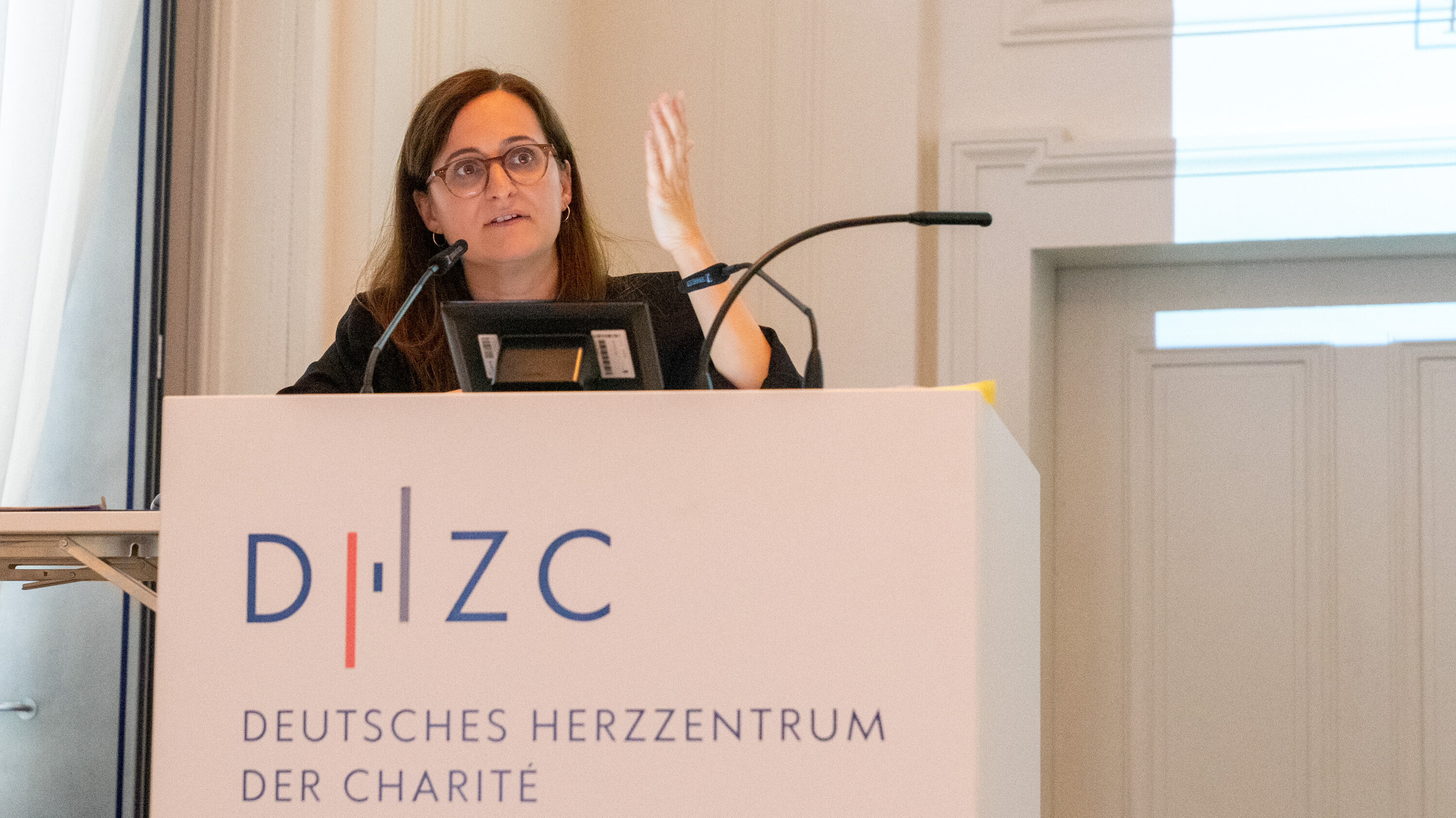Eine Person steht hinter einem Podium mit dem Schriftzug „Deutsches Herz-Zentrum der Charité“ und hält einen Vortrag. Sie gestikuliert und spricht, während auf einer Wand hinter ihr eine Präsentation angezeigt wird.