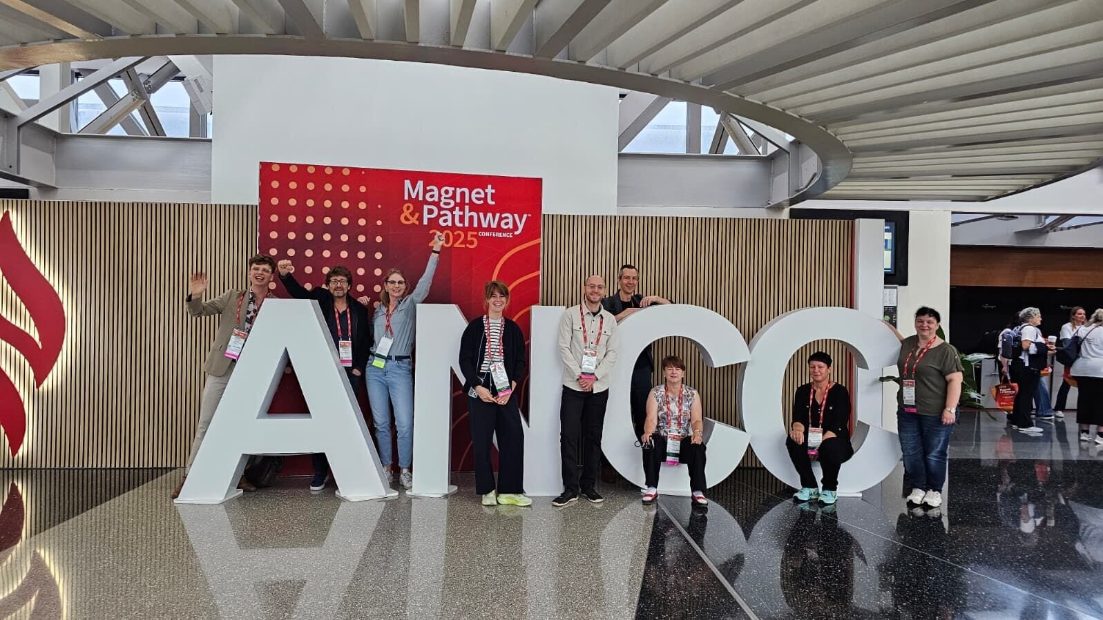 Eine Gruppe von Personen steht vor großen, weißen Buchstaben, die das Wort "AANCC" bilden. Im Hintergrund ist ein Banner mit dem Text "Magnet & Pathway Conference 2025" zu sehen. Die Teilnehmer sind gut gelaunt und posieren für das Foto.