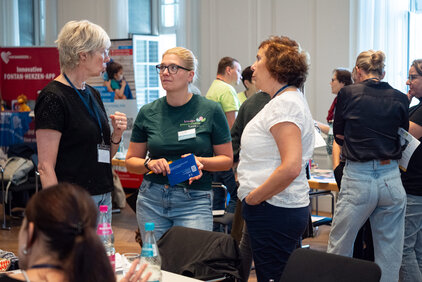 Drei Frauen stehen in einem Raum und unterhalten sich. Eine Frau trägt ein grünes T-Shirt und hält ein blaues Dokument in der Hand. Im Hintergrund sind weitere Personen und Tische zu sehen, die eine informelle Atmosphäre schaffen.