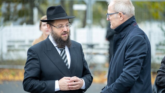 Zwei Männer führen ein Gespräch im Freien. Der Mann links trägt einen schwarzen Anzug und einen Hut, hat einen Bart und eine Brille. Der andere Mann steht rechts, trägt einen dunklen Mantel. Im Hintergrund sind Unschärfen von Personen und einer Umzäunung zu sehen.