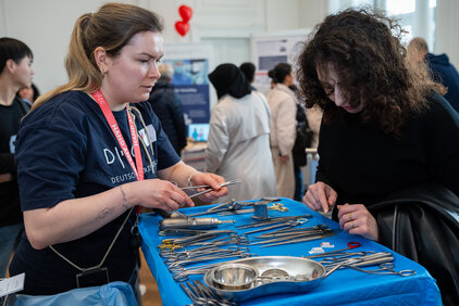 Eine Person zeigt einer anderen verschiedene chirurgische Instrumente auf einem Tisch. Beide Frauen sind in ein Gespräch vertieft. Im Hintergrund sind weitere Besucher zu sehen, die sich umsehen. Der Tisch ist ordentlich mit verschiedenen Werkzeugen und Geräten gedeckt.