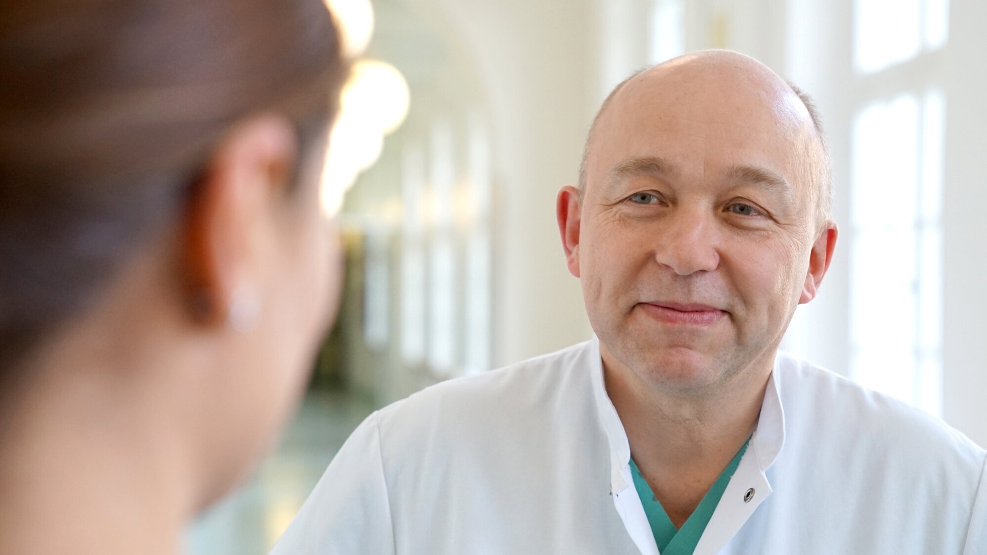 Ein lächelnder Arzt in einem weißen Kittel spricht mit einer Patientin. Der Hintergrund zeigt einen hellen, modernen Flur, der eine freundliche und einladende Atmosphäre schafft.