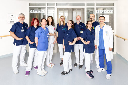 Eine Gruppe von 12 Personen in Krankenhauskleidung steht in einer hellen Flureingang. Sie lächeln und repräsentieren verschiedene medizinische Berufe. Im Hintergrund sind Glastüren mit der Aufschrift "STATION 45" sichtbar.
