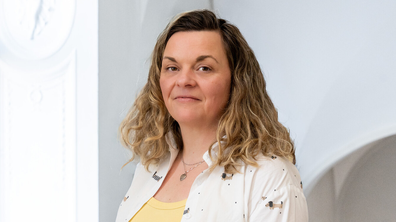 Porträt einer Frau mit lockigem, blonden Haar, die in einem hellen Raum steht. Sie trägt ein weißes Oberteil mit einem gelben Shirt darunter. Die Frau schaut freundlich, während sie leicht zur Seite blickt.
