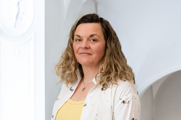 Porträt einer Frau mit lockigem, blonden Haar, die in einem hellen Raum steht. Sie trägt ein weißes Oberteil mit einem gelben Shirt darunter. Die Frau schaut freundlich, während sie leicht zur Seite blickt.