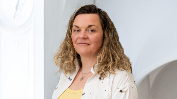 Porträt einer Frau mit lockigem, blonden Haar, die in einem hellen Raum steht. Sie trägt ein weißes Oberteil mit einem gelben Shirt darunter. Die Frau schaut freundlich, während sie leicht zur Seite blickt.