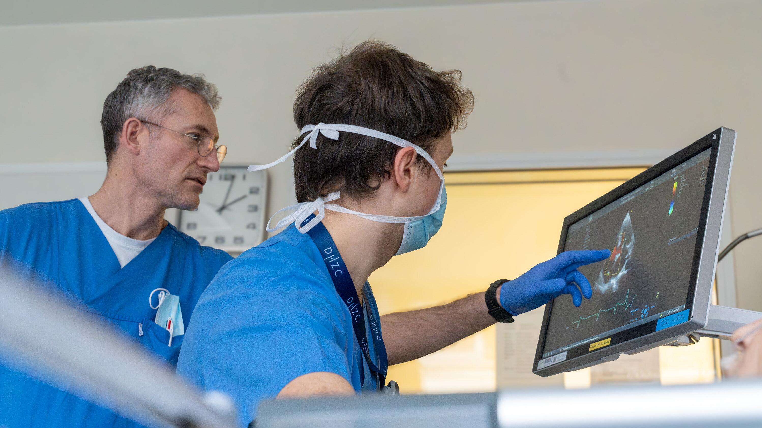 Zwei medizinische Fachkräfte in blauen Uniformen betrachten einen Monitor. Einer zeigt mit dem Finger auf den Bildschirm, während der andere aufmerksam zusieht. Der Monitor zeigt eine bildgebende Darstellung, wahrscheinlich von einem medizinischen Verfahren.