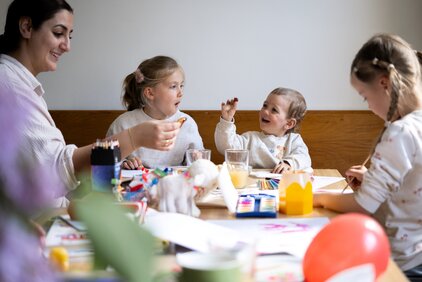 Eine Frau und drei Kinder sitzen an einem Tisch und malen. Die Kinder scheinen fröhlich zu sein, während sie mit Farben und Papier arbeiten. Es herrscht eine kreative und entspannte Stimmung. Es gibt verschiedene Malutensilien und Getränke auf dem Tisch.