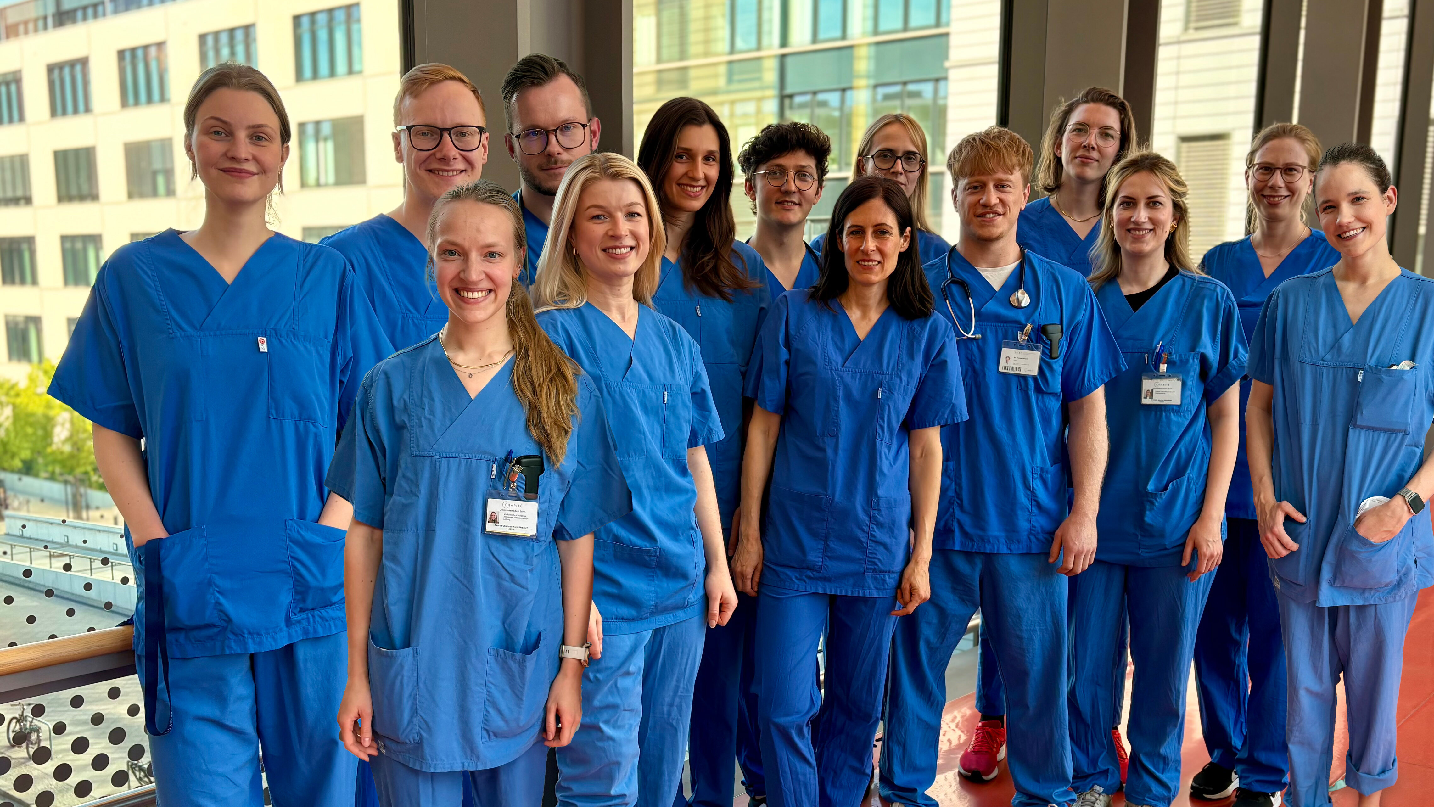 Eine Gruppe von 15 Personen in blauen Klinikoberteilen und -hosen steht lächelnd in einem Raum mit großen Fenstern. Im Hintergrund sind moderne Gebäude sichtbar. Die Szene vermittelt eine freundliche, professionelle Atmosphäre.