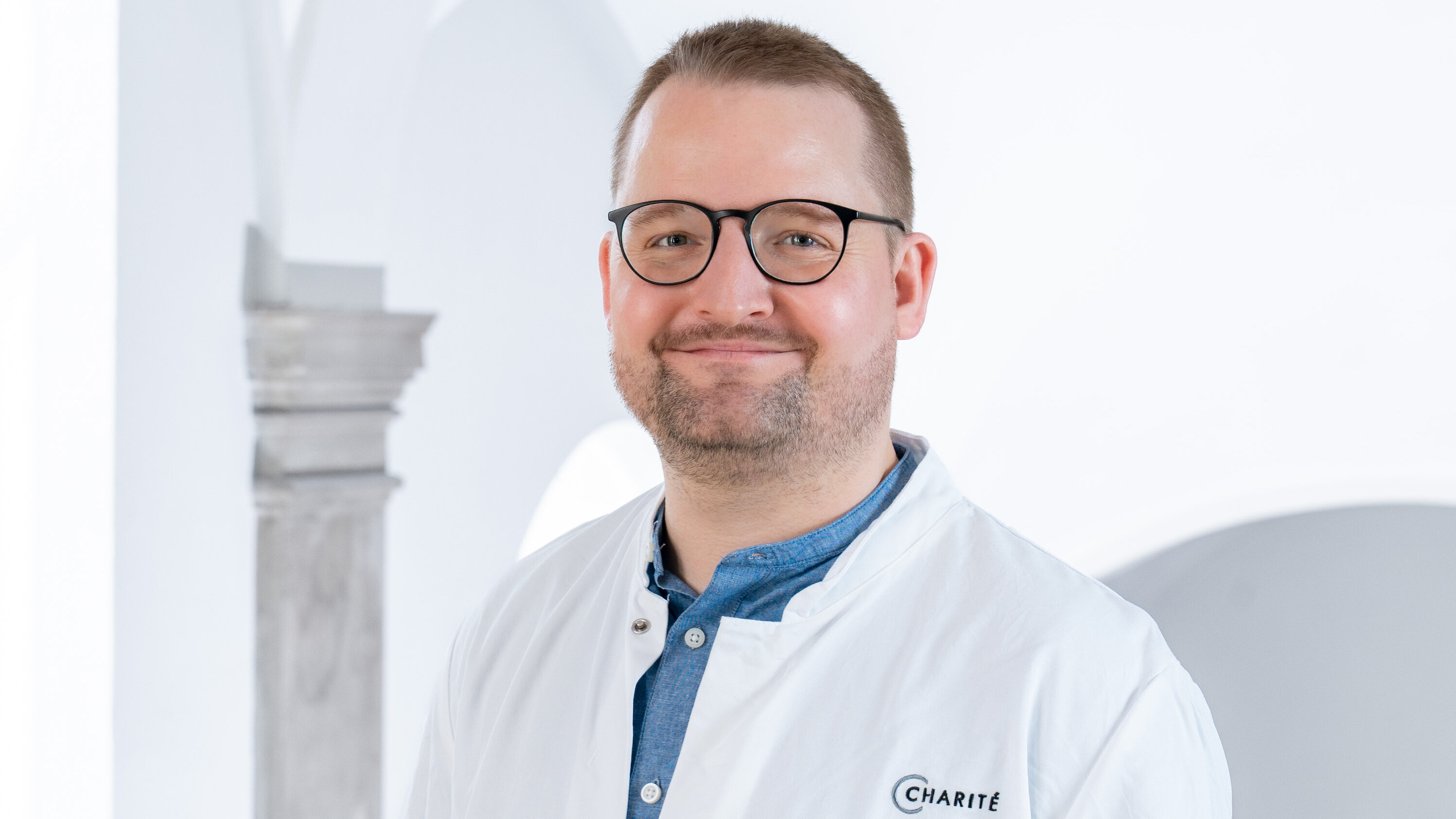 Ein lächelnder Arzt trägt einen weißen Kittel und eine Brille. Im Hintergrund sind helle Wände und architektonische Details zu sehen. Die Person strahlt Freundlichkeit und Professionalität aus.