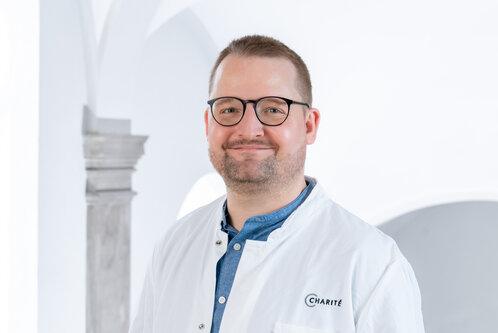 Ein lächelnder Arzt trägt einen weißen Kittel und eine Brille. Im Hintergrund sind helle Wände und architektonische Details zu sehen. Die Person strahlt Freundlichkeit und Professionalität aus.