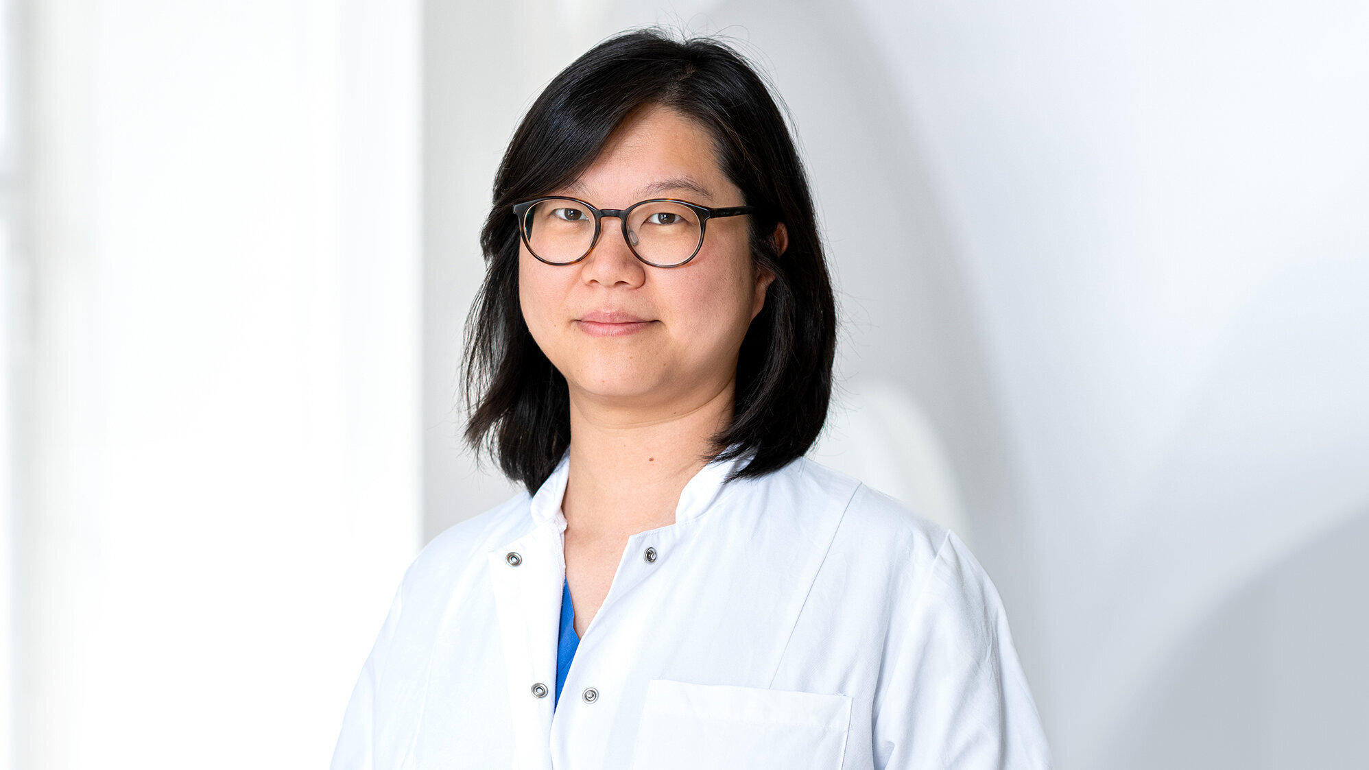 Eine Ärztin in einem weißen Kittel steht lächelnd vor einem hellen, neutralen Hintergrund. Sie trägt eine Brille und hat schwarzes, mittellanges Haar. Ihr Ausdruck vermittelt Professionalität und Freundlichkeit.