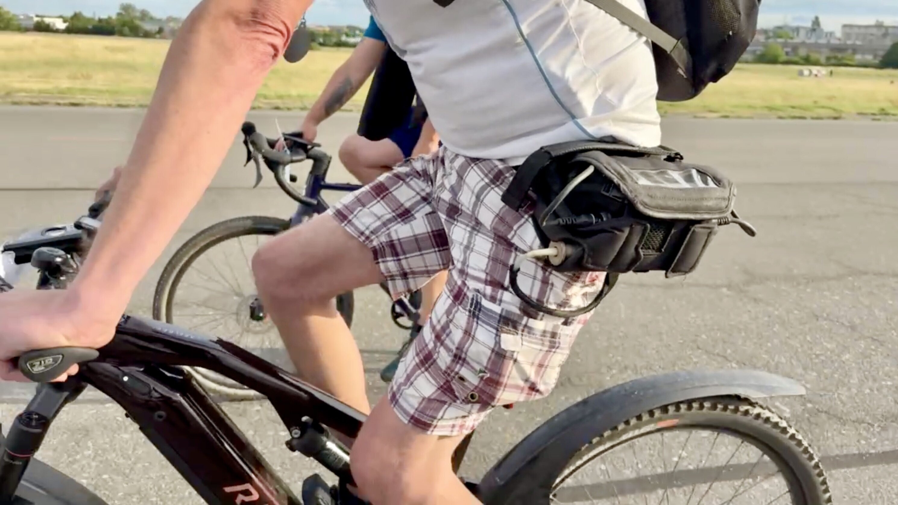 Zwei Personen fahren auf Fahrrädern. Im Vordergrund ist ein Mann zu sehen, der in einem weißen T-Shirt und karierten Shorts sitzt. Er trägt einen Rucksack und hat eine Tasche am Fahrrad. Der Hintergrund zeigt eine offene Fläche mit etwas Grün.