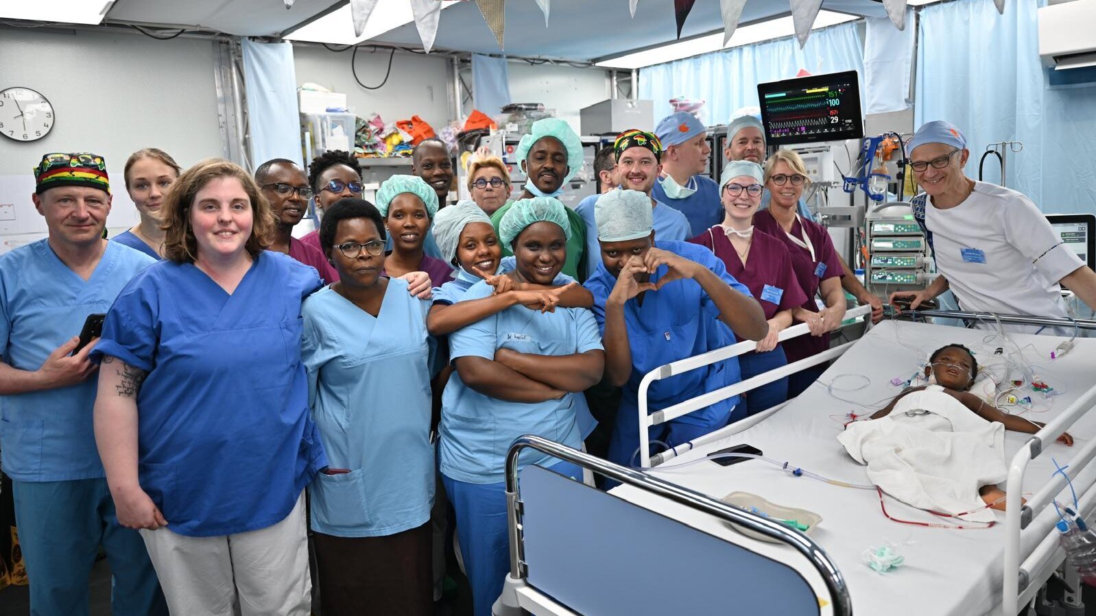 Eine Gruppe von medizinischem Personal, darunter Ärzte und Pflegekräfte in blauen und grünen Scrubs, steht lächelnd zusammen in einem Krankenhauszimmer, während ein kleines Kind auf einem Bett im Vordergrund liegt. Alle zeigen eine positive Einstellung und unterstützen den kleinen Patienten.