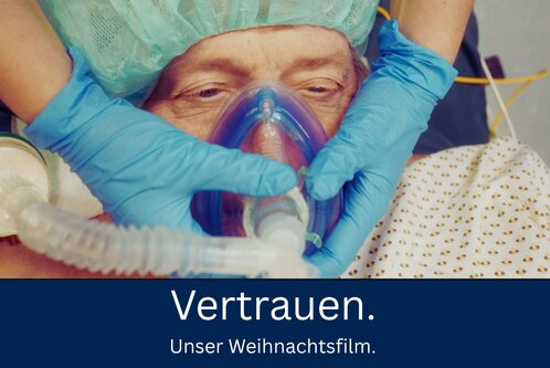 Nahaufnahme eines Patienten mit einer Atemmaske. Eine Pflegekraft mit blauen Handschuhen passt die Maske an. Im unteren Bildbereich steht der Text "Vertrauen. Unser Weihnachtsfilm." auf einem dunklen Hintergrund.
