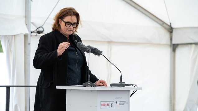 Eine Person steht an einem Rednerpult mit Mikrofon und spricht. Sie trägt einen schwarzen Mantel und hat braune, lockige Haare. Im Hintergrund ist ein Zelt sichtbar. Auf dem Pult sind Logos und Schriftzüge zu sehen, die auf Sponsoren hinweisen.
