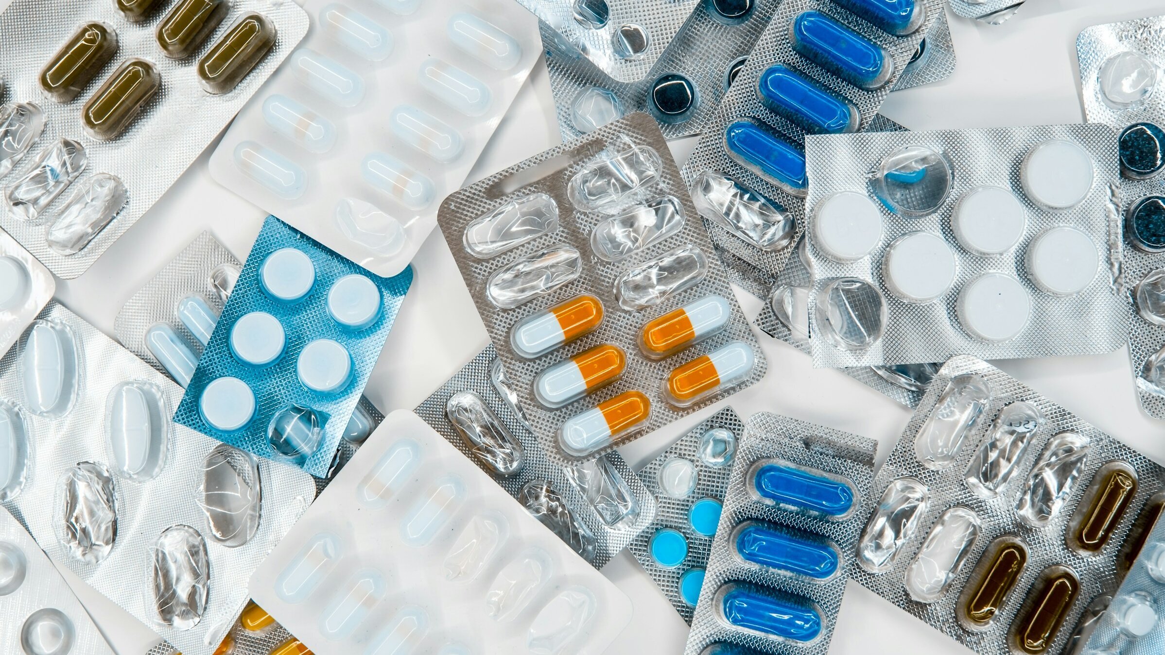Das Bild zeigt eine Vielzahl von Medikamentenverpackungen, darunter verschiedene Tabletten und Kapseln in unterschiedlichen Farben und Formen. Die Verpackungen sind durcheinander auf dem Hintergrund angeordnet, was eine große Auswahl an pharmazeutischen Produkten darstellt.