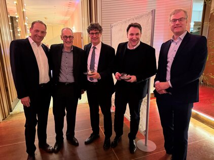 Fünf Männer stehen nebeneinander in einem festlich dekorierten Raum. Sie halten jeweils einen Preis oder eine Auszeichnung in den Händen und lächeln zur Kamera. Die Atmosphäre wirkt feierlich und erfreulich.