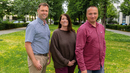 Drei Personen stehen auf einer Wiese umgeben von Bäumen. Sie lächeln freundlich und scheinen miteinander zu interagieren. Im Hintergrund sind unscharfe Gebäude und ein Weg zu sehen. Die Atmosphäre ist entspannt und freundlich.