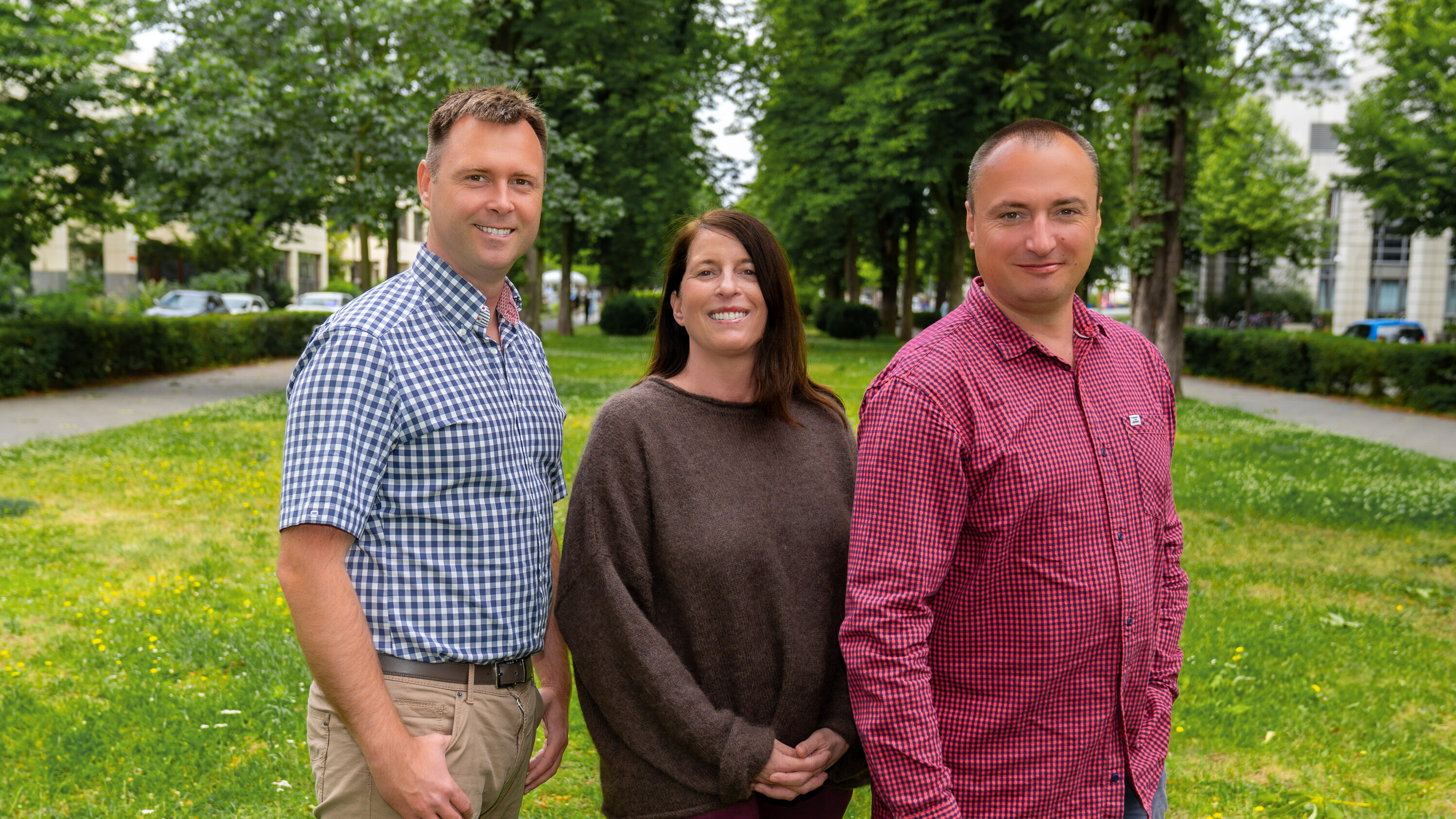 Drei Personen stehen auf einer Wiese umgeben von Bäumen. Sie lächeln freundlich und scheinen miteinander zu interagieren. Im Hintergrund sind unscharfe Gebäude und ein Weg zu sehen. Die Atmosphäre ist entspannt und freundlich.