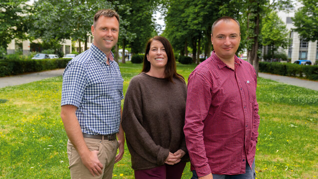 Drei Personen stehen auf einer Wiese umgeben von Bäumen. Sie lächeln freundlich und scheinen miteinander zu interagieren. Im Hintergrund sind unscharfe Gebäude und ein Weg zu sehen. Die Atmosphäre ist entspannt und freundlich.